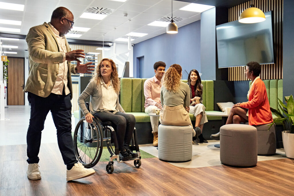 Full length view of small group sitting together and talking as mature man and woman walk and wheel past, face to face and exchanging ideas. ADA