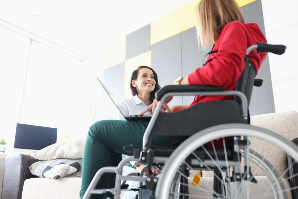 Disabled woman in wheelchair in communication with smiling friend with laptop on her lap. Psychological assistance to people with disabilities concept.