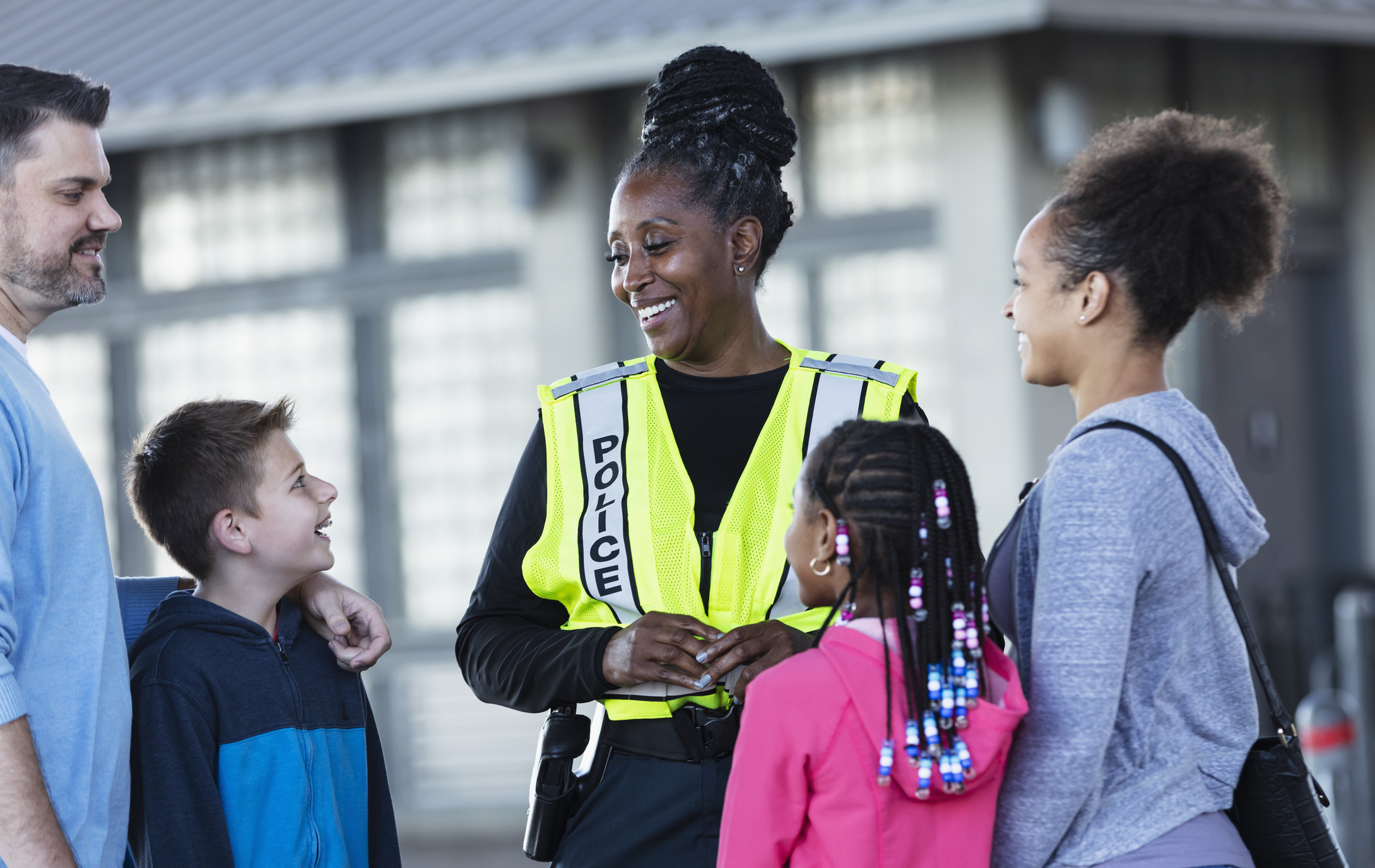 An African-American policewoman conversing with a multiracial group of parents and children outdoors. Everyone is smiling.