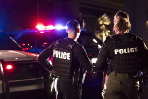 Rear view of two multiracial law enforcement officers standing side by side in front of their police vehicles with emergency lights on. They are working at night in a city. The policewoman is mixed race, African-American, Asian and Hispanic, in her 40s. Her partner is a young Hispanic man in his 20s.