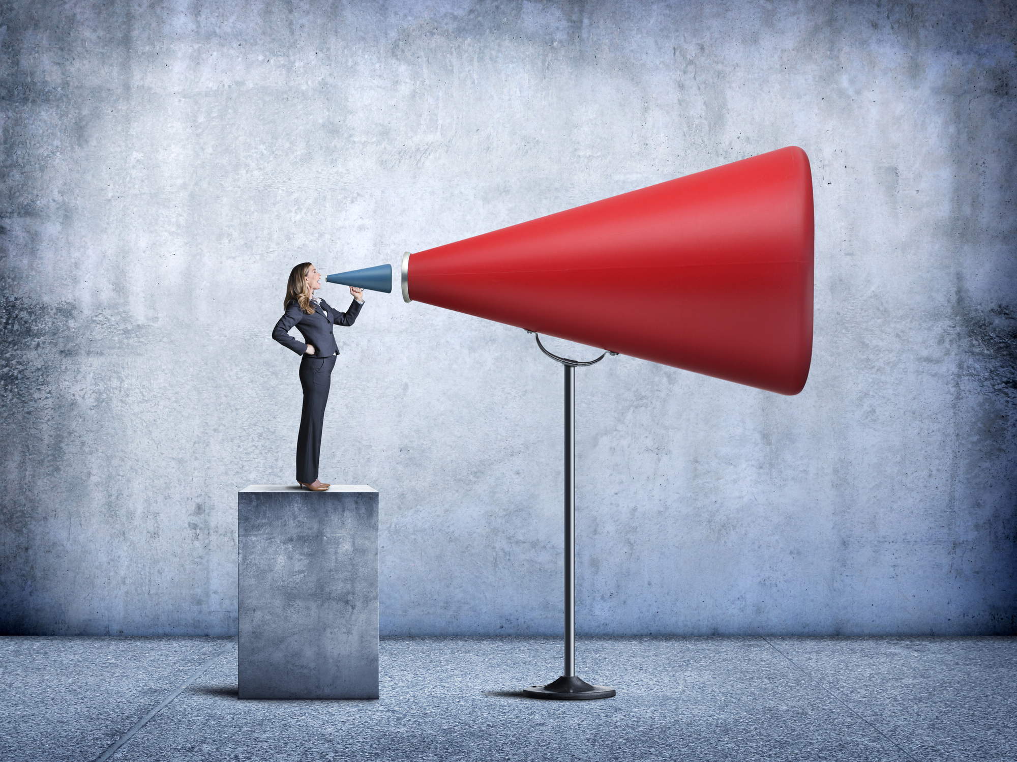 A businesswoman stands on a concrete pedestal as she uses a blue megaphone to shout into a much larger red megaphone in order to amplify her message.