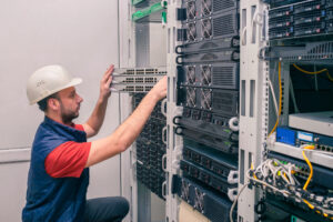 A man installs new equipment in a modern data center. An engineer in a white helmet mounts a stack of switches in a rack. The technician works in the server room.
